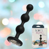 A black beaded silicone plug featuring a jeweled base, labeled as "Booty Bling Silver Silicone Anal Beads" by Doc Johnson, is displayed beside its packaging. The background showcases a pastel blur that complements the elegance of these fashionable anal beads.